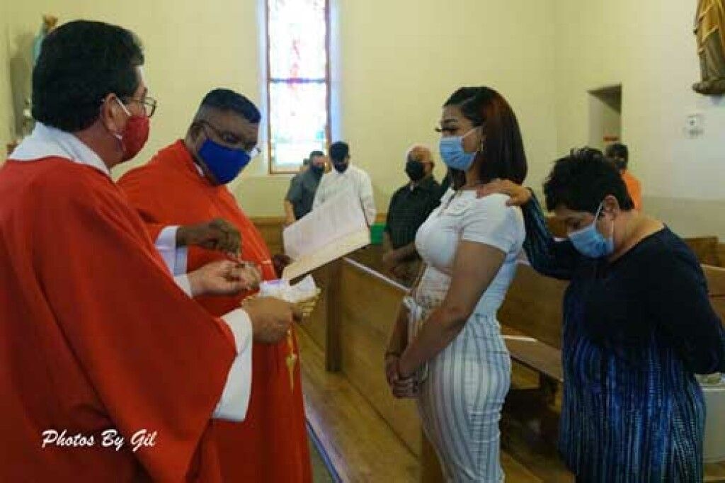 A priest in red robes conducts a church ceremony with two women wearing masks.