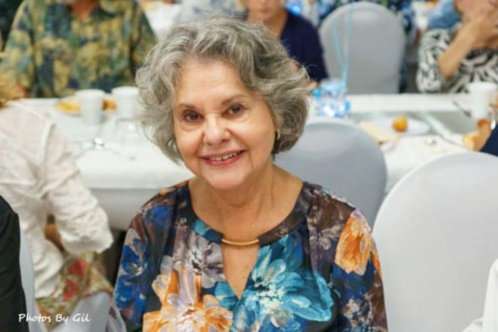 Elderly woman with gray hair smiles warmly at a banquet, wearing a floral blouse. 