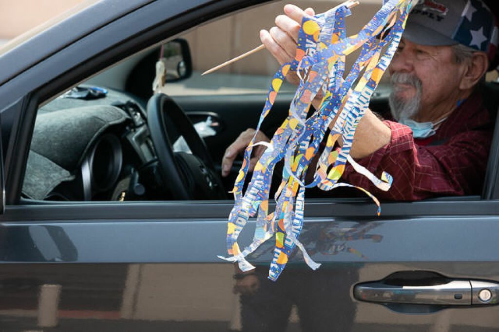 A man with a beard, wearing a cap, sits in a car with the window down, holding colorful paper streamers. 