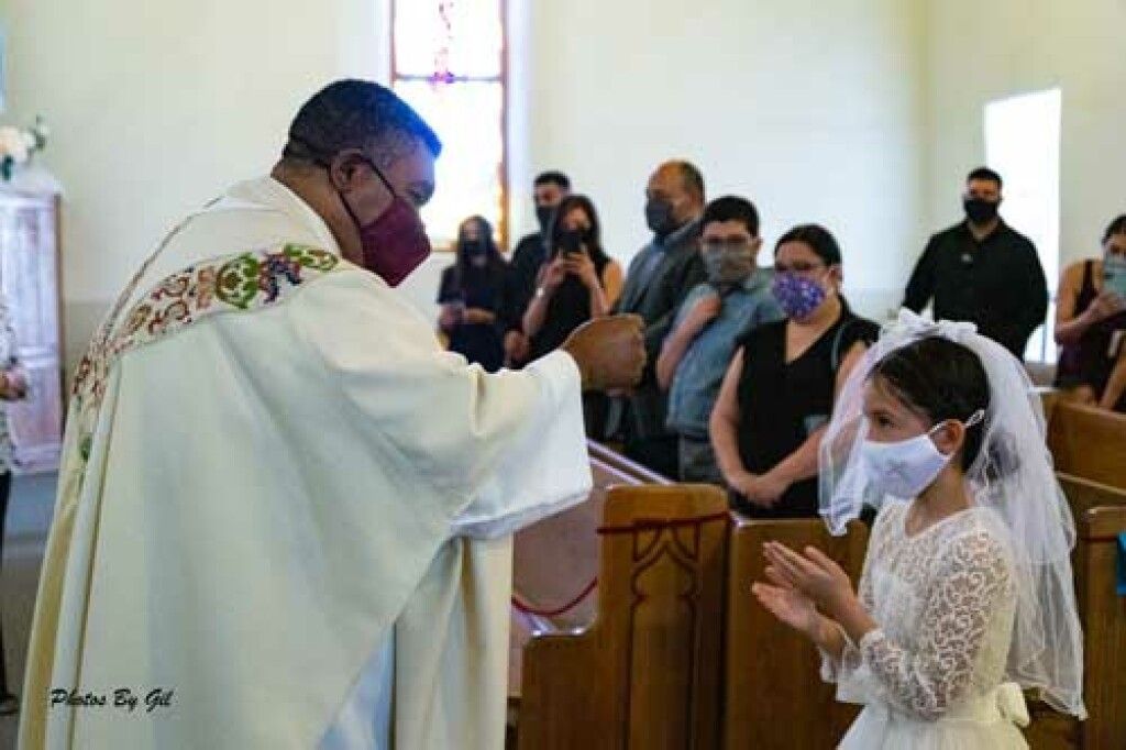 A priest in a white robe blesses a young girl in a white dress and veil, both wearing masks.