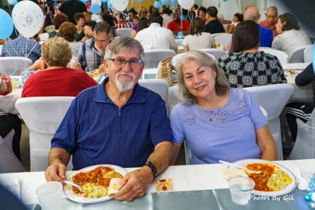 Elderly couple smiling at a communal dining event, seated at a table with plates of food. 