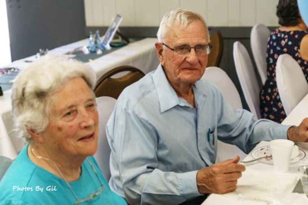 An elderly man and woman seated at a dining table, wearing casual attire. 