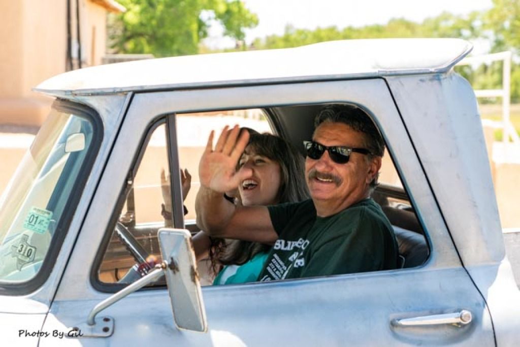 A smiling man in sunglasses and a woman wave from an old silver truck. 