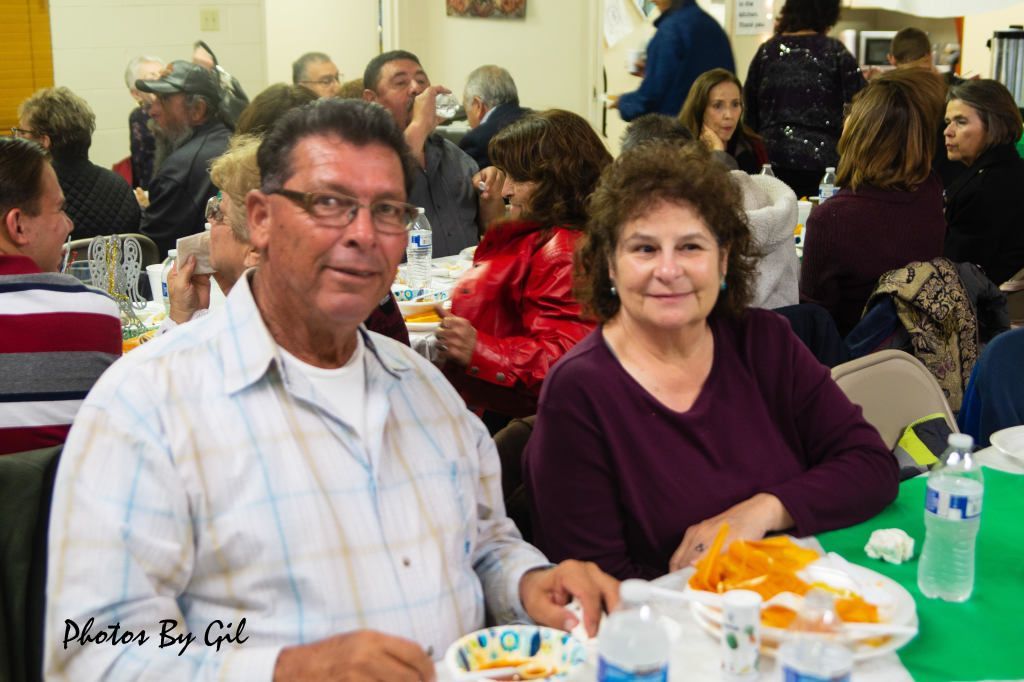 A smiling man and woman sit at a table with plates of food at a crowded gathering. 