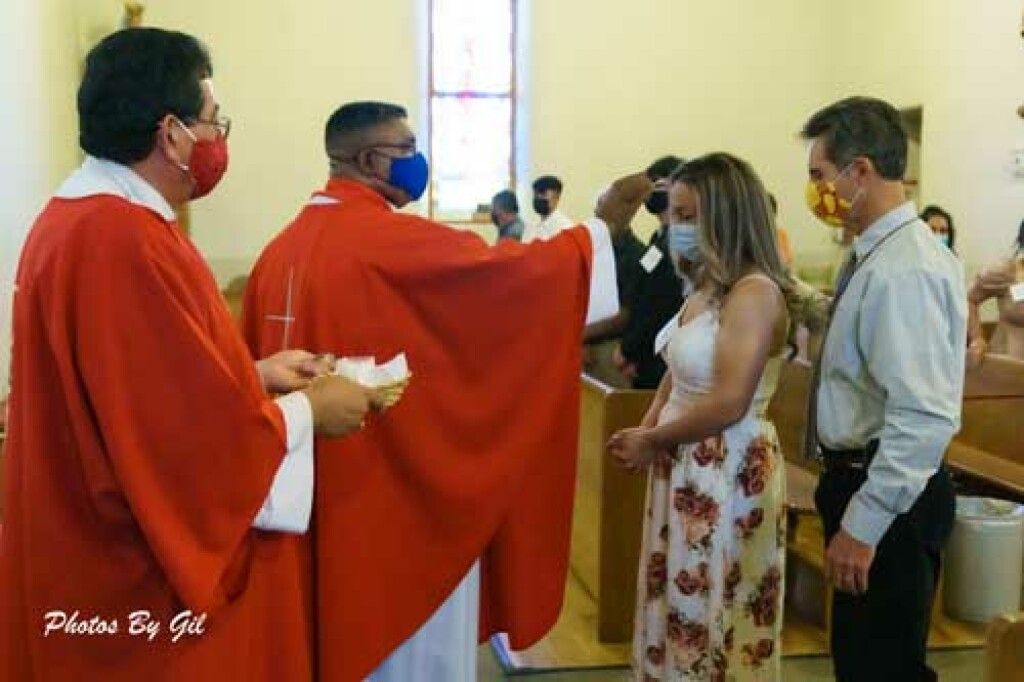 A priest in red robes performs a blessing on a woman in a floral dress, while another priest holds a container. 