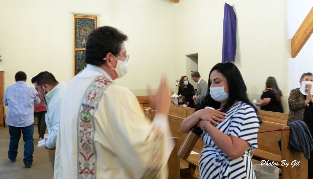 A masked priest gestures a blessing to a woman in a church aisle.
