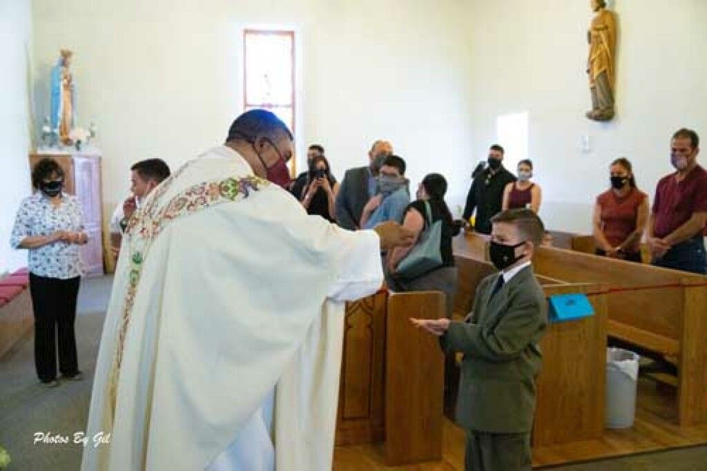 A priest in white robes, wearing a mask, performs a blessing for a masked man in a suit in a church.
