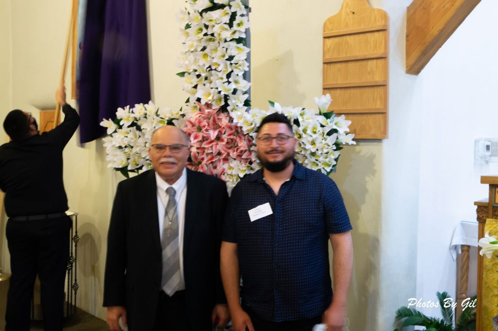 Two men stand smiling in front of a large floral cross made of white and pink flowers. 