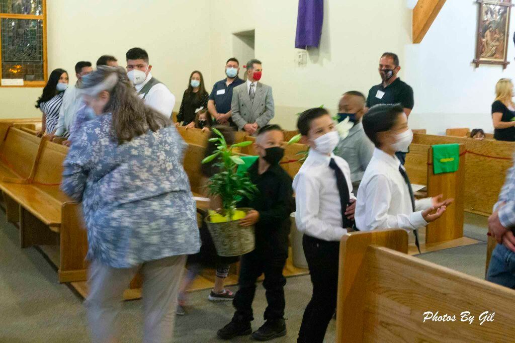 Children wearing masks and formal attire carry plants in a brightly lit church.