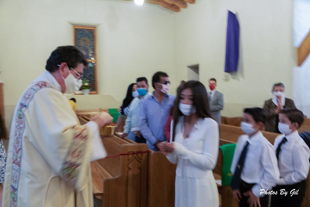 A priest in a decorated robe gives Communion to a masked girl in white.