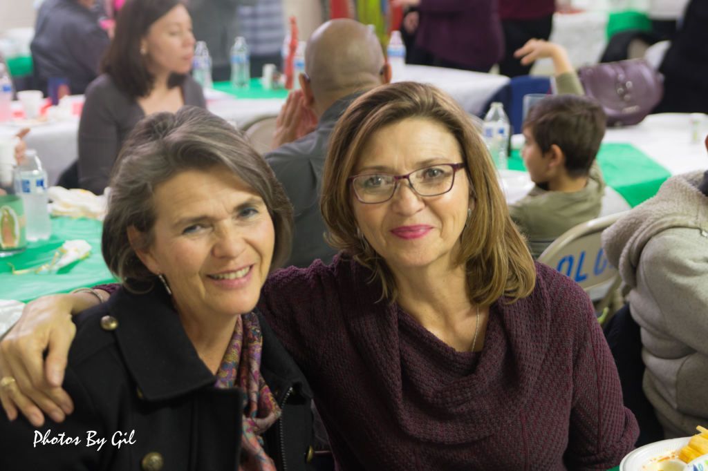 Two women smiling and sitting closely in a lively social setting.