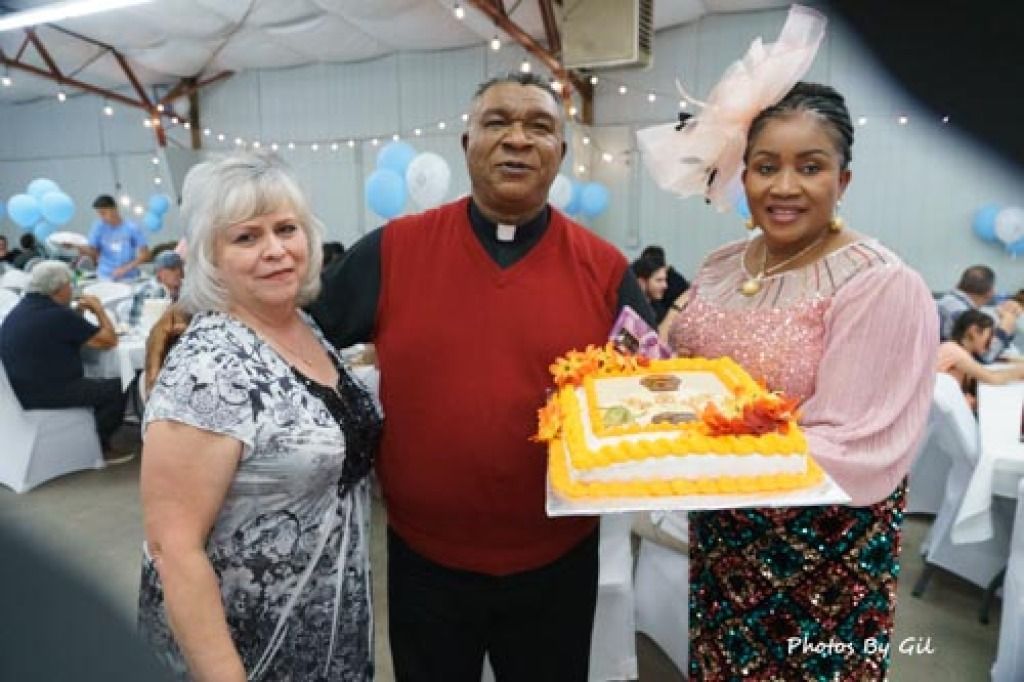 Three people smiling at a party, holding a decorated cake with orange frosting and floral designs.
