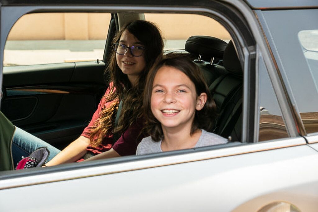 Two smiling children sit in the backseat of a car, visible through the open window. 