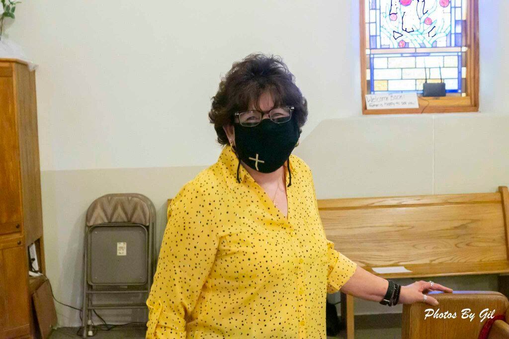 A person in a yellow polka-dot shirt and black mask with a cross symbol stands indoors, smiling.