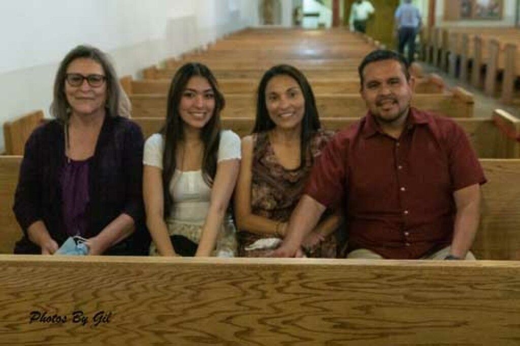 A group of four people sit together on a wooden pew, smiling warmly. 