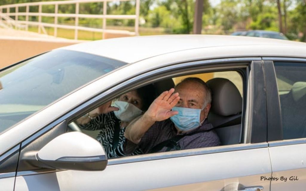 An elderly couple wearing masks waves from inside a silver car.