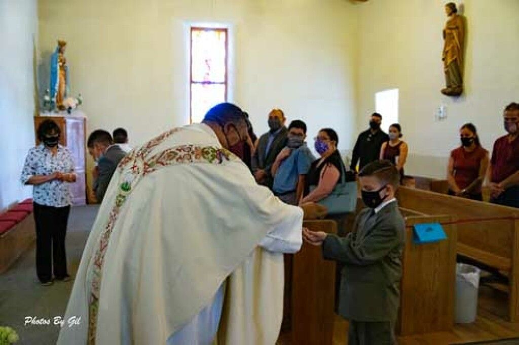 A priest in white robes, wearing a mask, performs a blessing for a masked man in a suit in a church.
