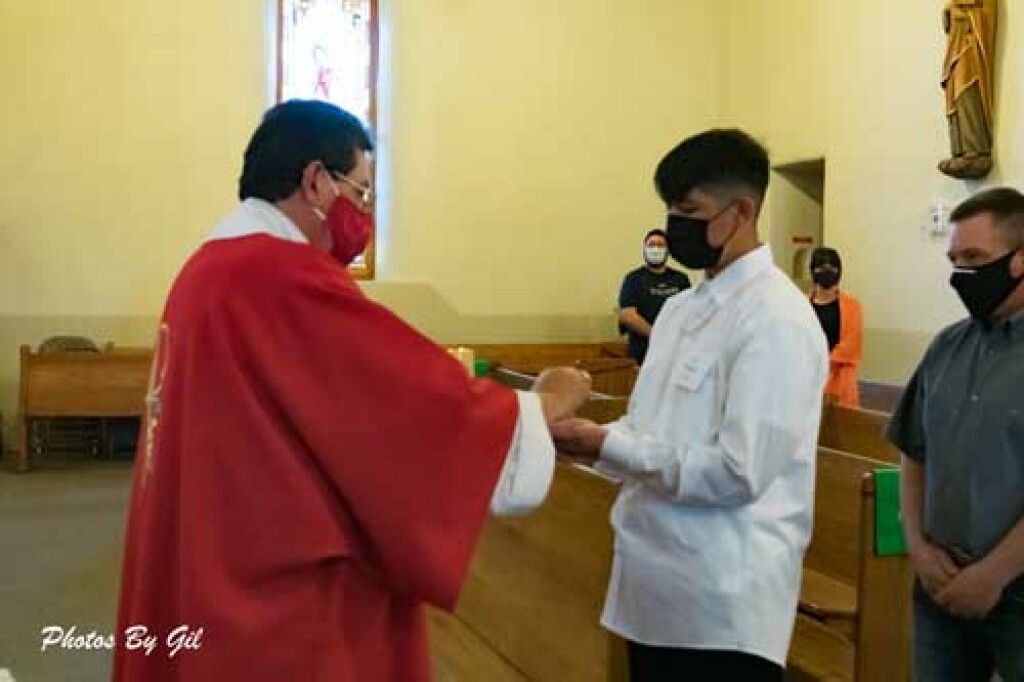 A priest in a red robe and mask is giving communion to a masked young man in a church. 