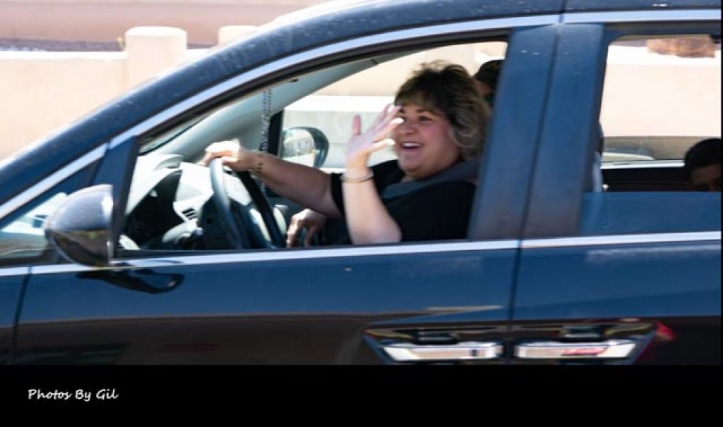 A woman in a black car joyfully waves from the driver’s seat.