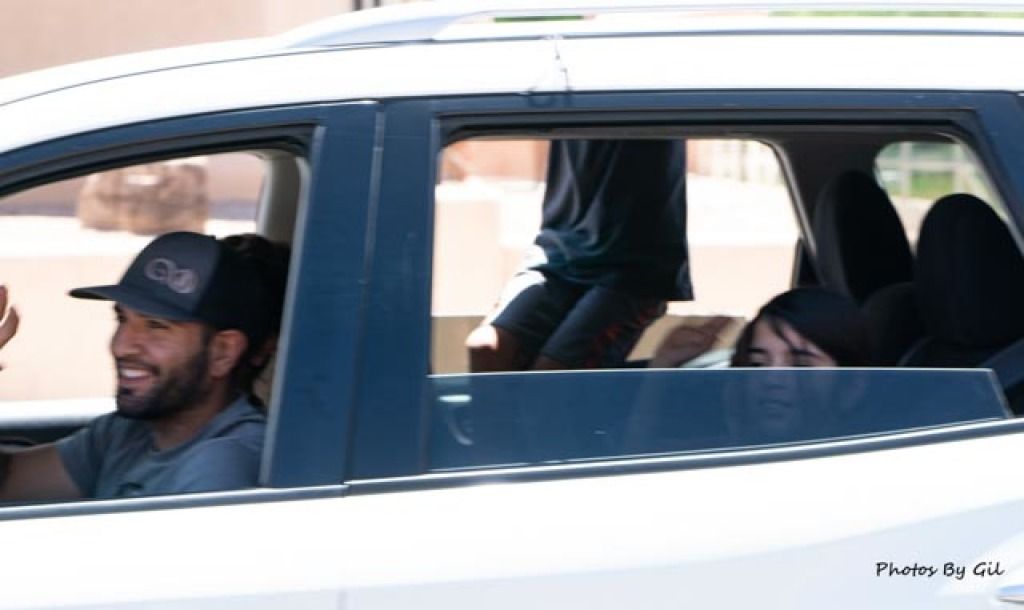 A man in a cap drives a white car, smiling and waving. 