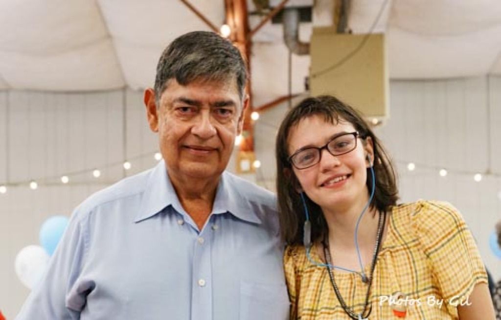 Elderly man in a blue shirt and a young woman in a yellow plaid dress smile warmly indoors. 
