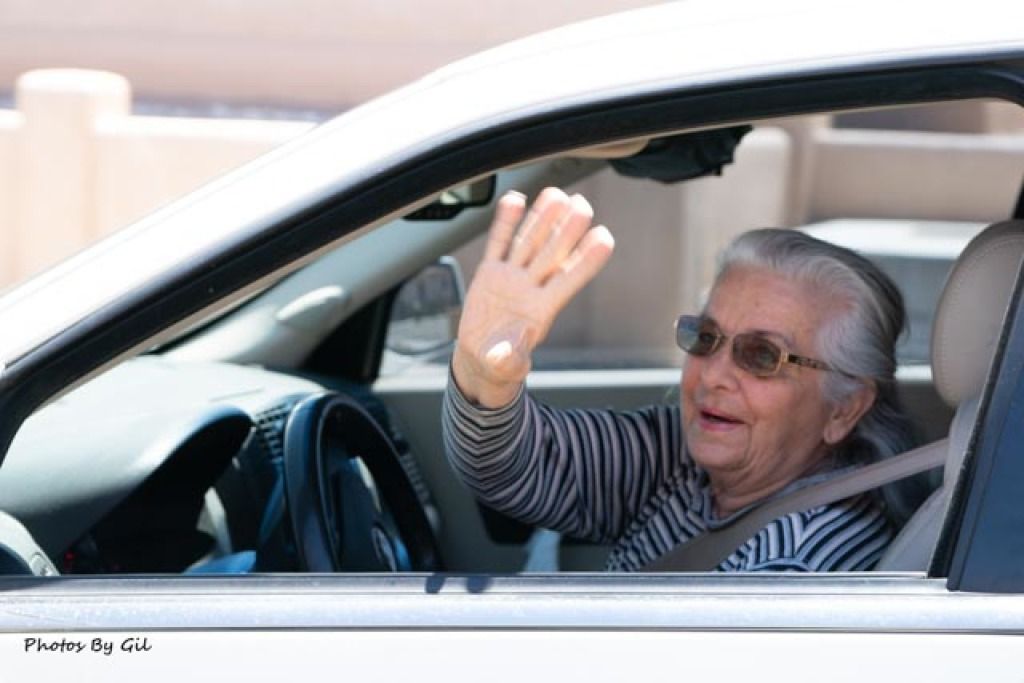 An elderly woman with gray hair and sunglasses waves cheerfully from the driver's seat of a car.