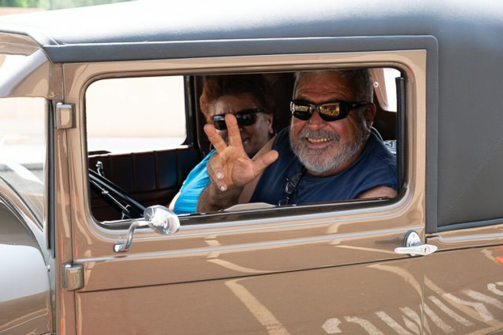 Smiling man in sunglasses flashes a peace sign from vintage car window, with woman beside him. 