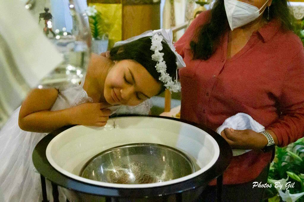 A young girl in a white dress and lace headband, smiling, leans over a baptismal font.