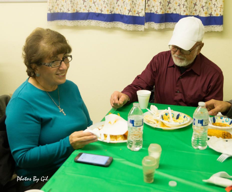 A smiling woman in glasses and a teal sweater sits at a table with a man in a white cap and maroon shirt.