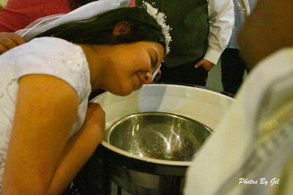 Smiling girl in a white dress and tiara leans over a baptismal font during a ceremony.