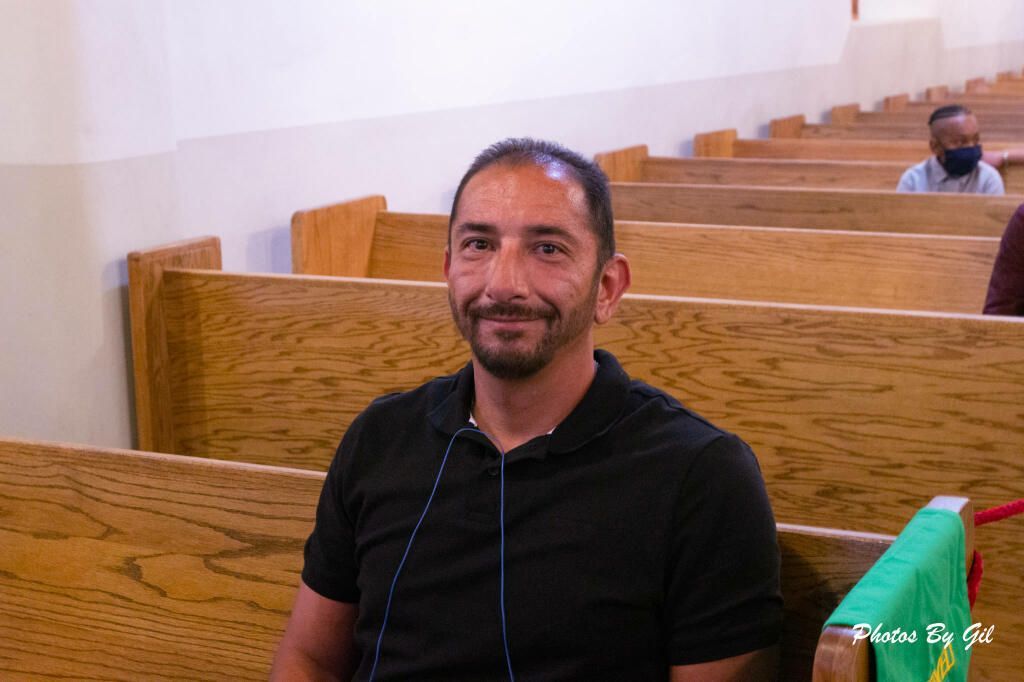 A man with a slight smile in a black shirt sits in a wooden pew, possibly in a church.