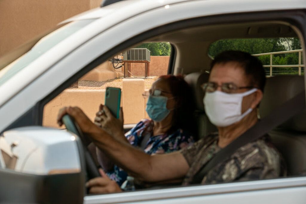 A man and woman sit in a parked car, both wearing face masks.