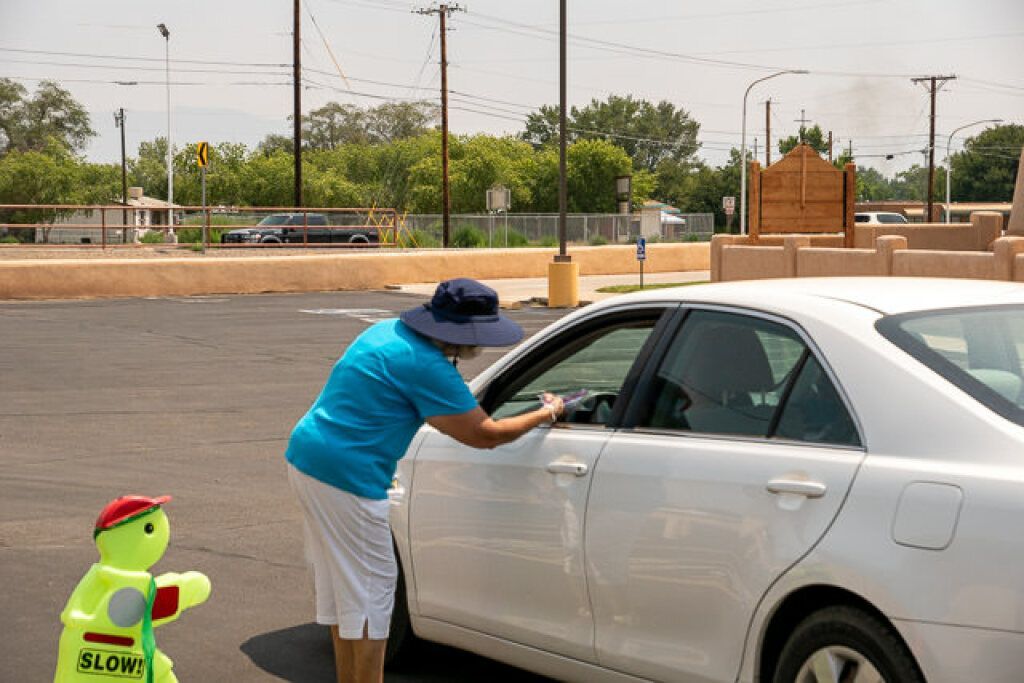 A person wearing a blue shirt and hat leans into a white car window in a parking lot.