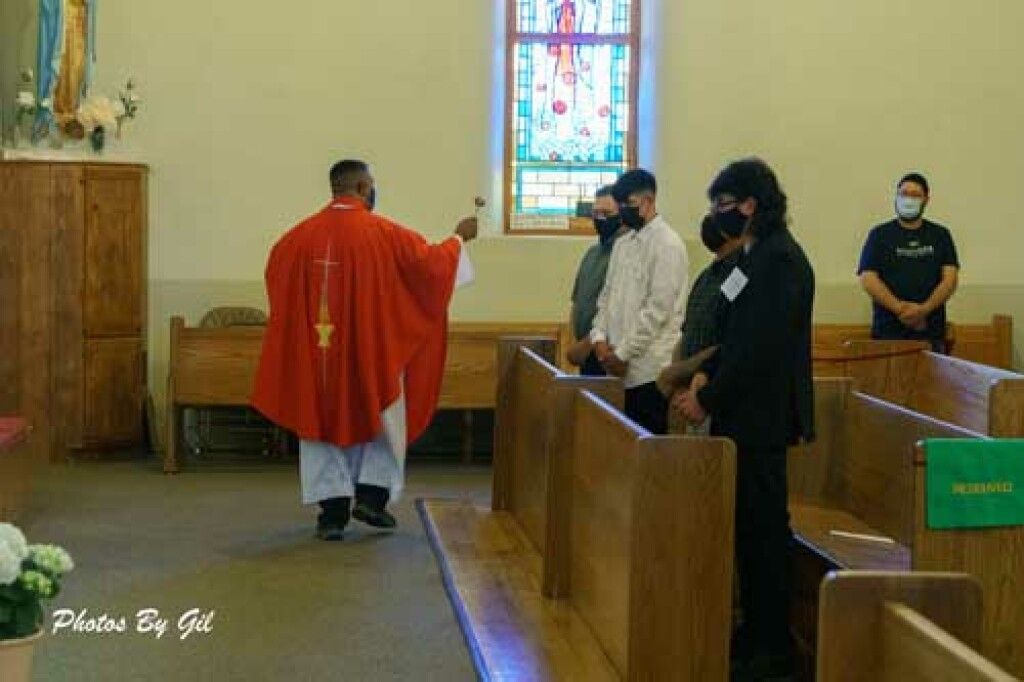 A priest in red vestments blesses four masked men inside a church with wooden pews.