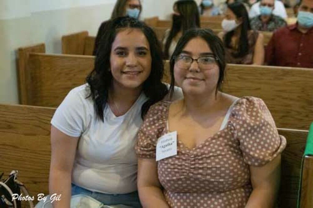 Two women sit closely on wooden pews, smiling warmly. 