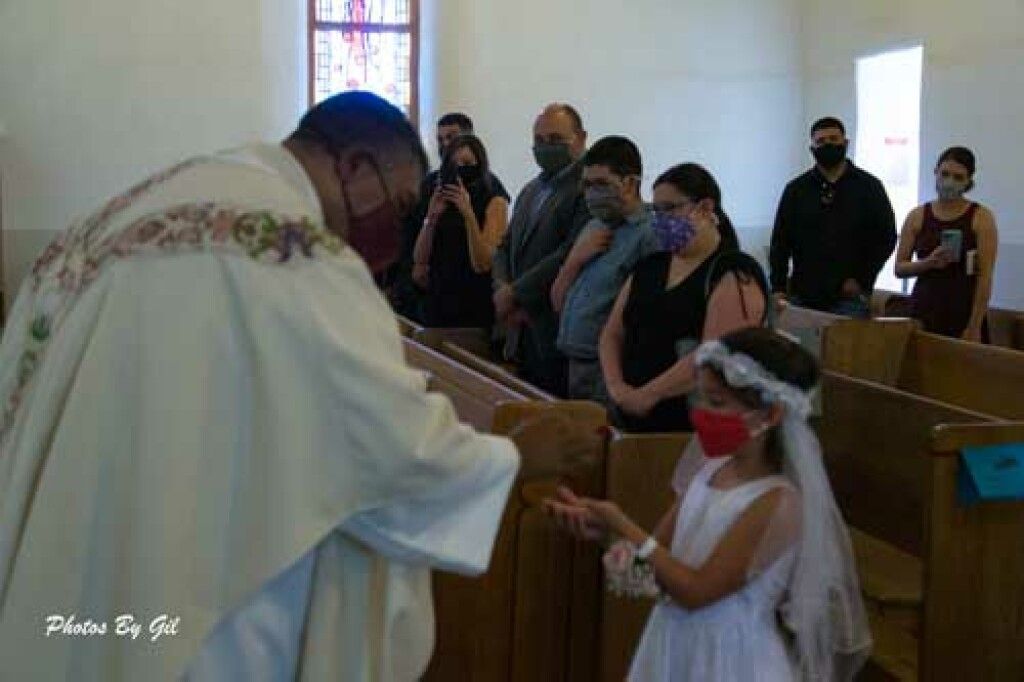 A priest hands communion to a young girl in a white dress and veil at a church.