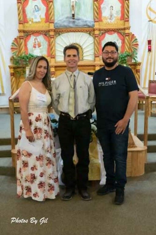 Three people stand together in a church with a colorful altar in the background.