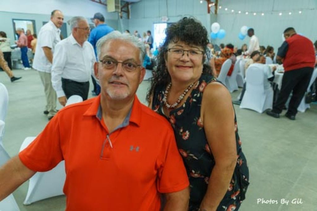 A man in a red shirt and a woman in a floral blouse smile at the camera in a lively banquet hall. 