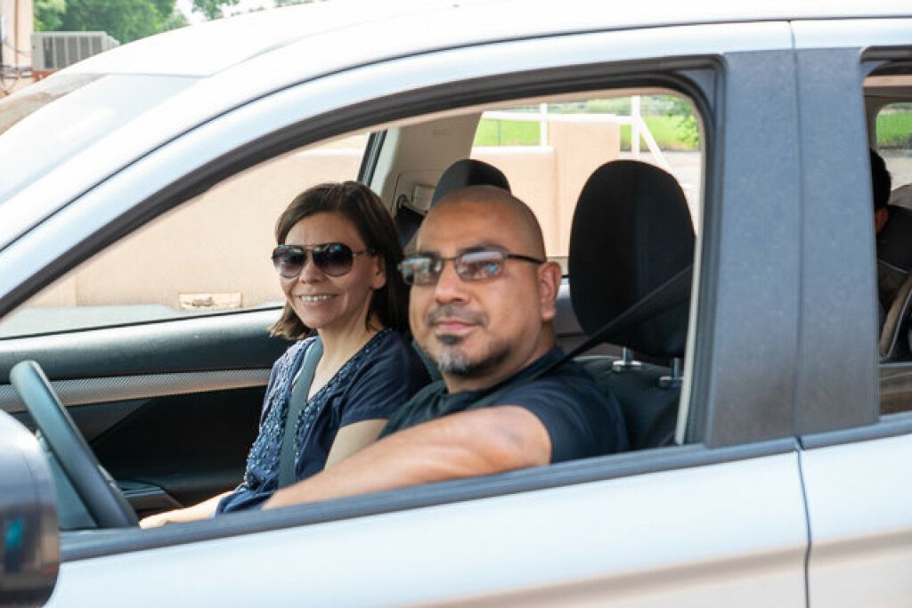 A man and woman sit in a parked car, both wearing sunglasses and smiling. 