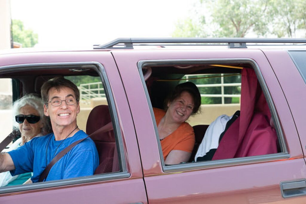 A man in blue and an elderly woman in sunglasses are in the front seat of a red SUV, both smiling.