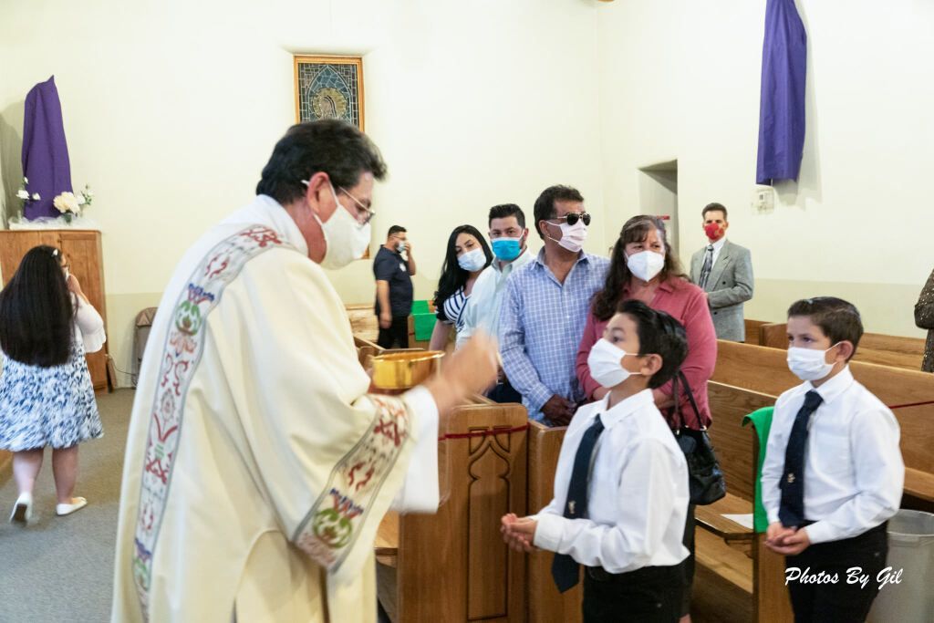 A priest in a decorated robe gives Holy Communion to two masked boys in white shirts and ties.