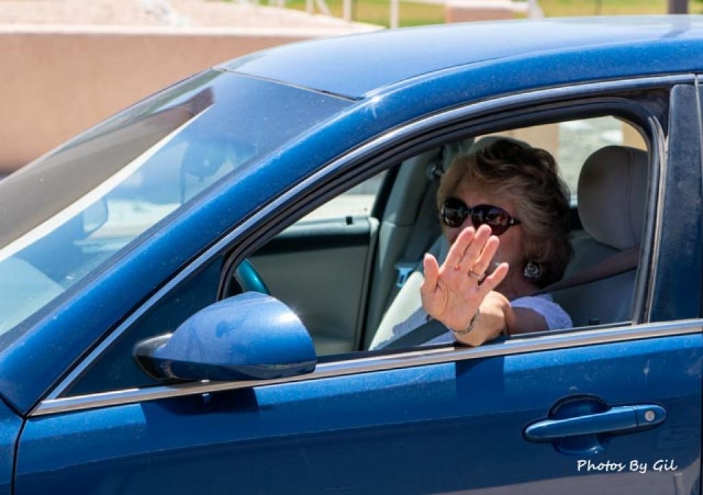 Woman inside a blue car, wearing sunglasses and gesturing with her hand out the window as if to stop or greet. 