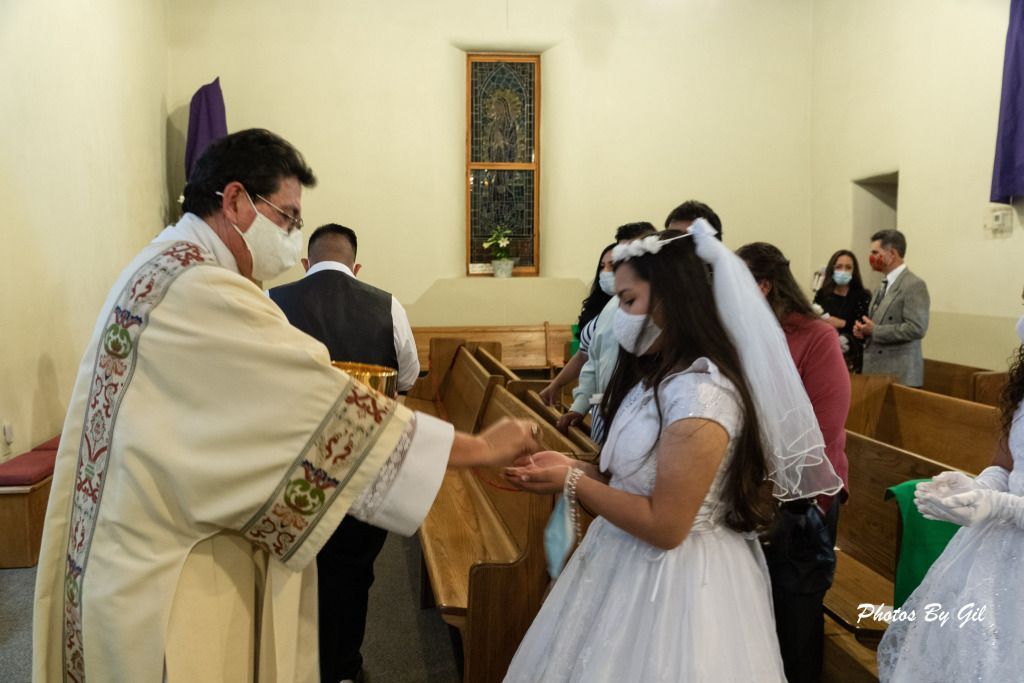 A priest in a decorated robe gives communion to a young girl in a white dress and veil. 