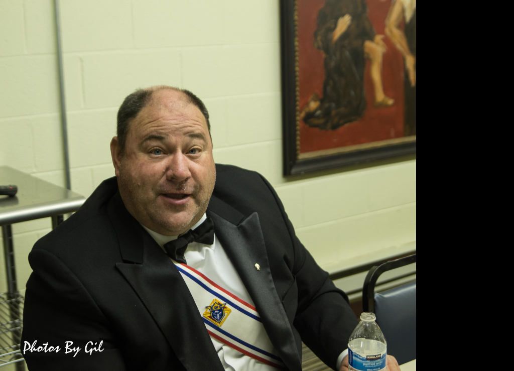 A man in a tuxedo and medal ribbon sits indoors, holding a water bottle.