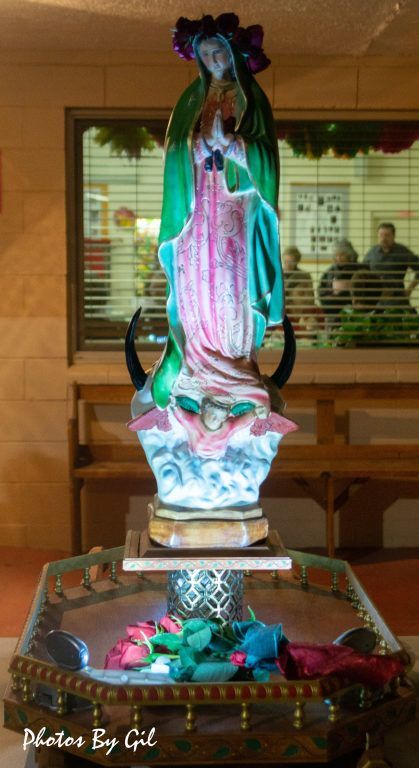 A statue of the Virgin Mary, draped in a green and pink robe, stands illuminated in a room. 