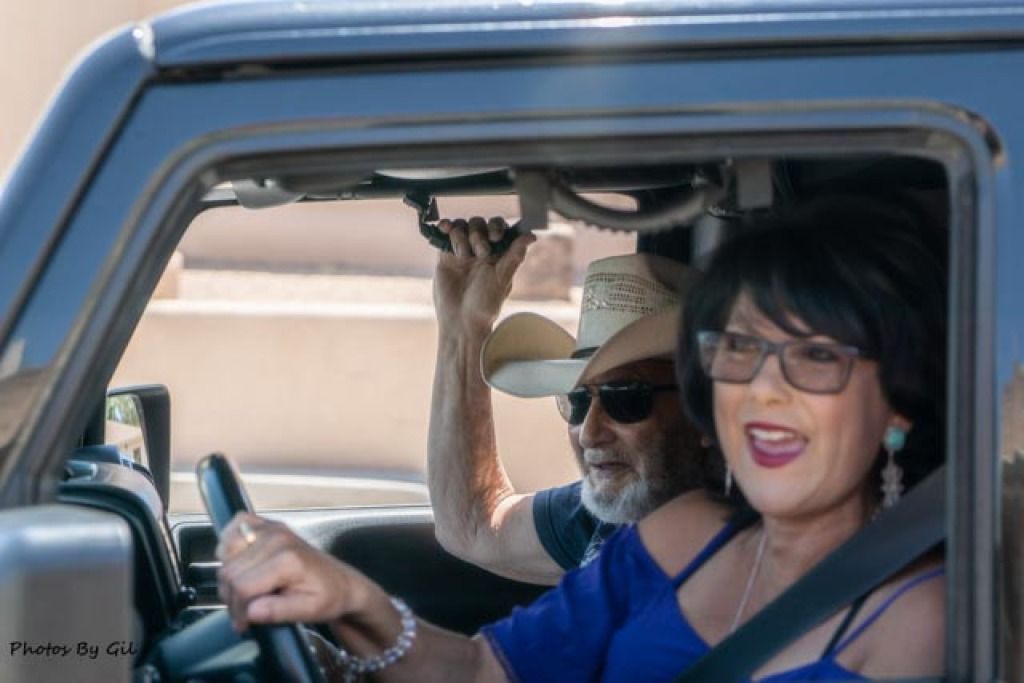 A woman and an older man in sunglasses and a straw hat are joyfully driving in a car with open windows.