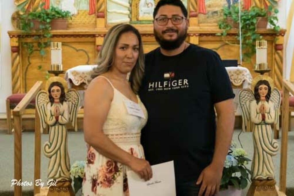 A couple stands smiling in a decorated church with wooden angel statues. 
