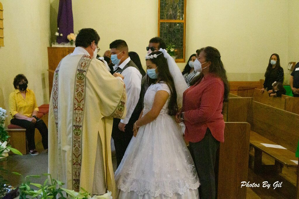 A priest in ornate robes blesses a girl in a white dress during a church ceremony.