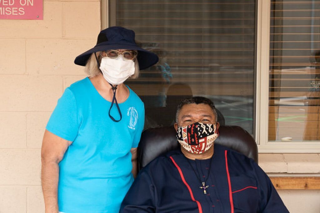 A woman in a blue shirt and sun hat stands beside a seated man wearing a patterned face mask and cross.