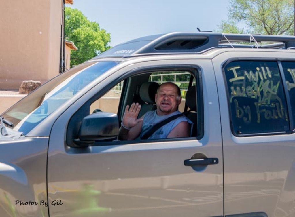 A man in a sleeveless shirt waves cheerfully from a parked SUV.